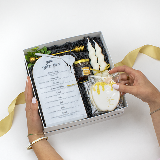 Hands holding a Rosh Hashana Gift Box with Simanim Plaque, set of twisted taper candles, honey jar, gold honey dipper, Shana Tova matches and apple shaped sugar cookies in white box