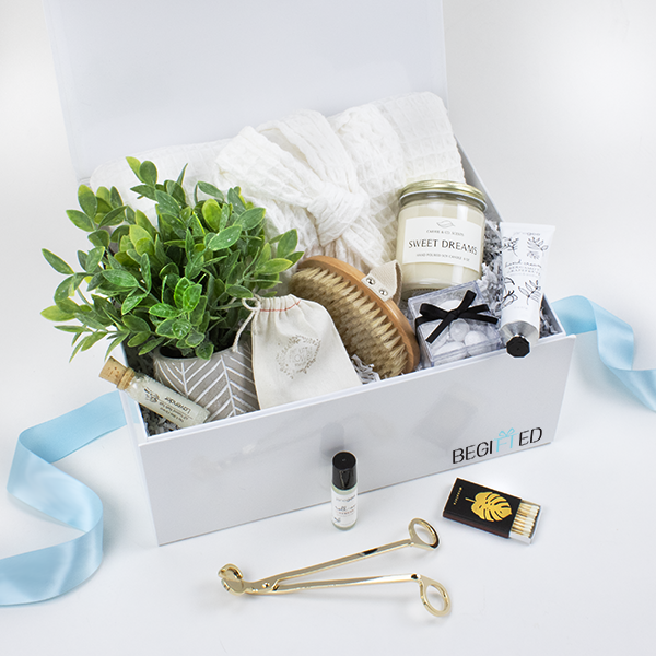 The Blissful Spa Gift Box featuring a white magnetic gift box, waffle weave robe, sweet dreams scented candles, bath salts, hand cream, bath brush, candle wick cutter, matches with gold embossed box, lavender scented body roll on, and faux plant in stone holder.
