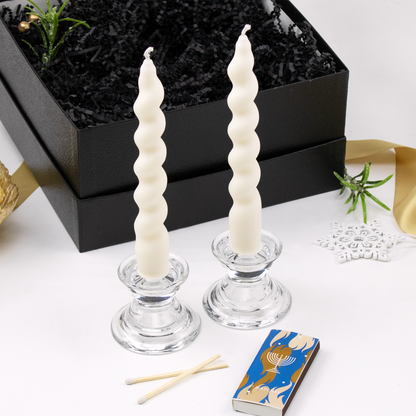 Two twisted tapered candles in glass holders and box of matches with menorah printed on it on a white surface.