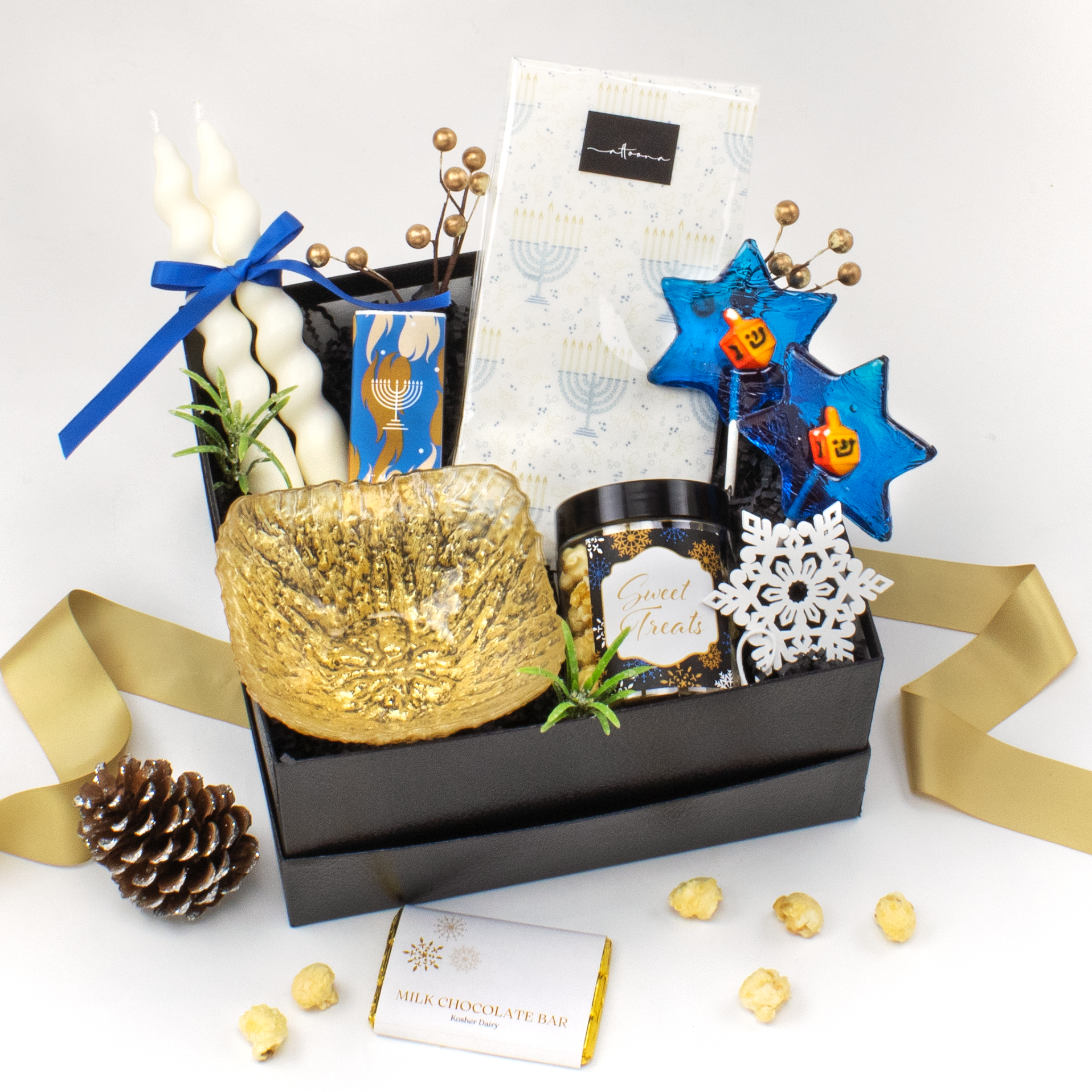 Gift box with tapered candles, menorah printed guest towels, gold etched snack bowl, matches, chocolates and snowflake ornament on white background