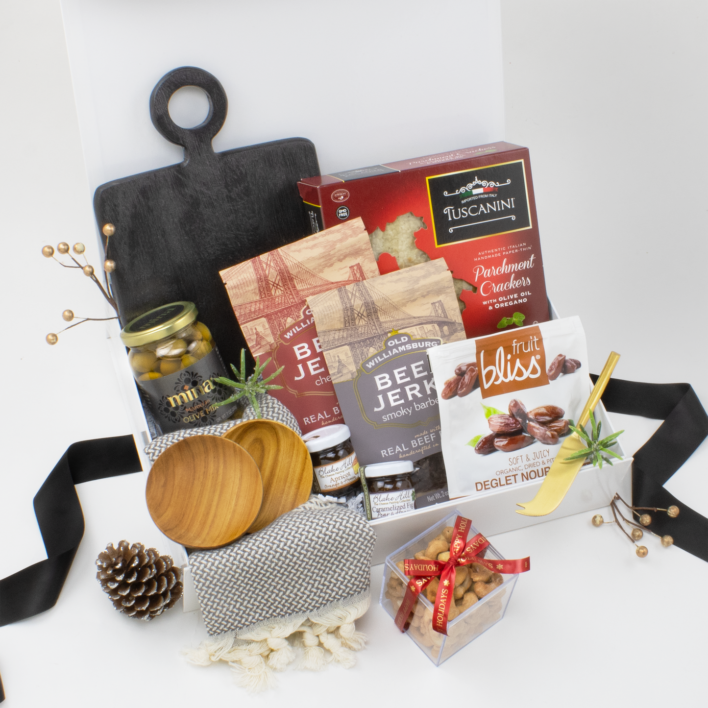 Assorted gourmet food items including a wood serving board, acacia wood snack bowls, cotton kitchen towel and gold spreading knife in a white gift box on white background