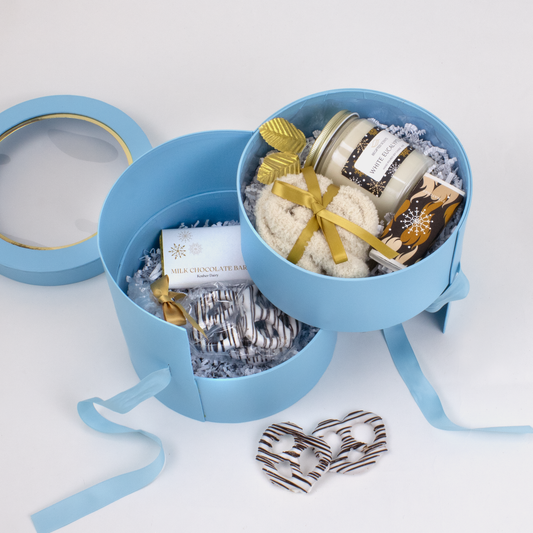 Open gift set with a candle, cozy socks, matchbox, pretzels and chocolate in a blue round 2-layer gift box on a white background