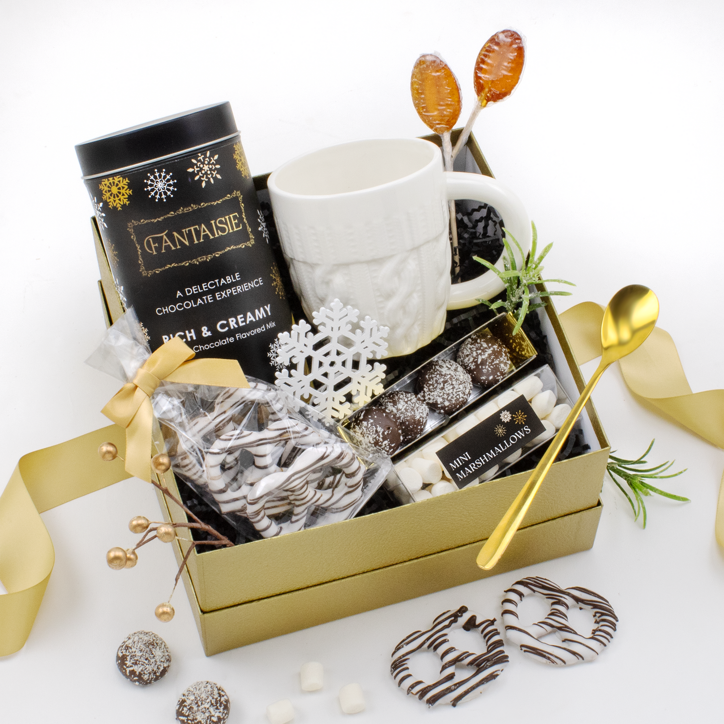 Gift basket with hot chocolate, a mug, marshmallows, gingerbread lollipop spoons, gold long handled spoon, pretzels, chocolate truffles on a white background