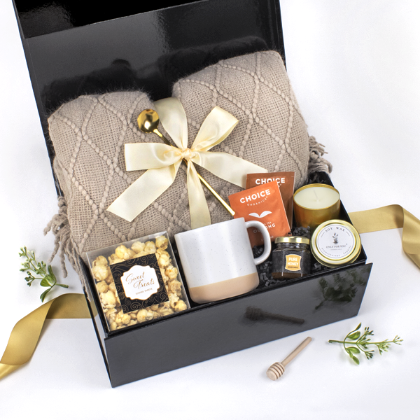 The Cozy Home Gift Set with a throw blanket, stoneware mug, tea bags, scented candle, honey jar and wood dipper and flavored popcorn treats. 